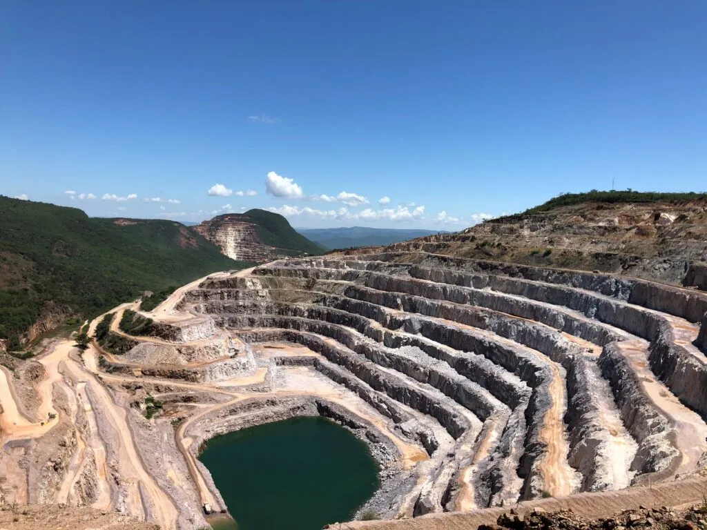 Mina a céu aberto com níveis em espiral, estrada de acesso e lago no fundo, cercada por montanhas verdes.